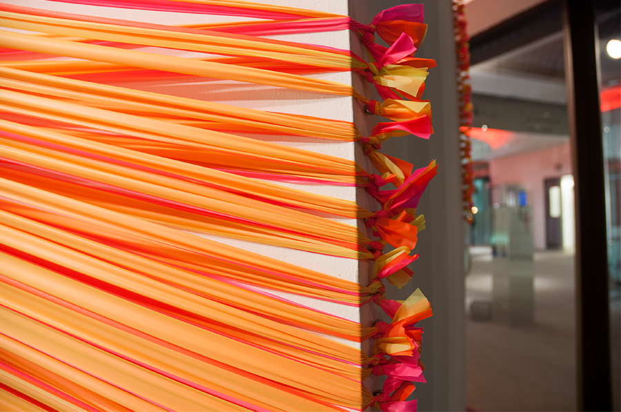 A detail image of florescent pink and yellow strands of flagging tape stretching around the rear corners of an architectural column, terminating in a series of eye hooks.
