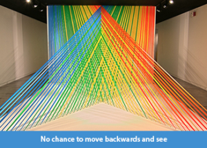 A photo of a site-specific art installation made of linear strands of multi-colored flagging tape arranged in planes that intersect each other in space terminating at a 17 foot long pedestal.