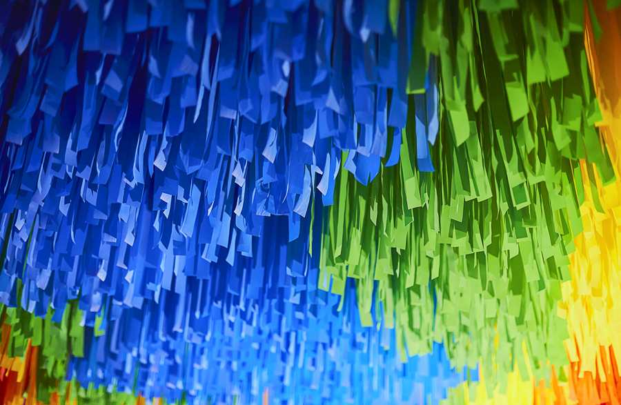 A detail image of a rainbow-hued, large-scale installation at the Laguna Art Museum in Laguna Beach, California that is suspended from the ceiling, comprised of colorful flagging tape.
