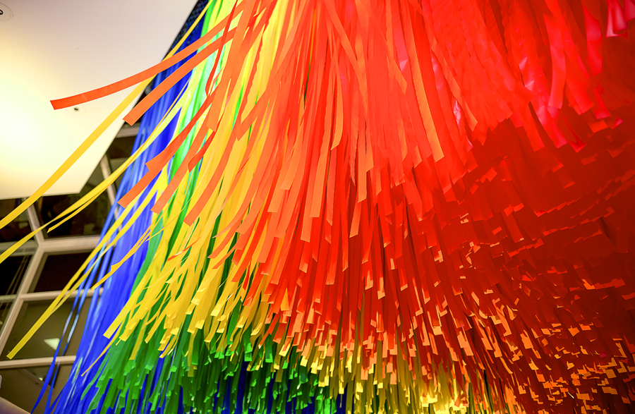 A detail image of a rainbow-hued, suspended flagging tape installation, taken from the floor, looking up into the plane of color.